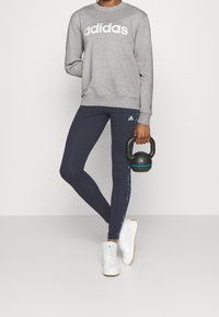 Person som bär en grå Adidas sweatshirt, marinblå Adidas leggings och vita sneakers, håller en svart kettlebell vid sin sida mot en enkel bakgrund.