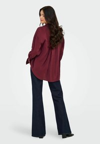 Donna con lunghi capelli ondulati e scuri, che indossa una maglietta larga bordeaux e jeans svasati scuri, in piedi con le mani nelle tasche, mentre guarda dietro di sé.