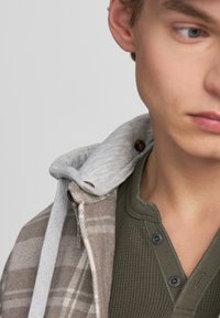 Kapturowana kurtka w kratę w miękkim beżu i szarości z guzikami; nałożona na teksturowaną oliwkowo-zieloną koszulę henley z dekoltem w serek i czarnymi guzikami.