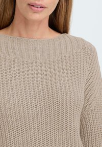 Beige pletený svetr s texturovaným vzorem, širokým výstřihem a žebrovanými detaily na ramenou. Měkká, pohodlná tkanina s volným střihem.