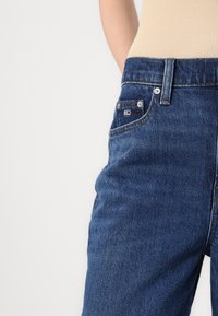 Osoba nosi plave traperice visokog struka s vidljivim prednjim džepom i malom oznakom logotipa, uparene s bež rebrastim topom.