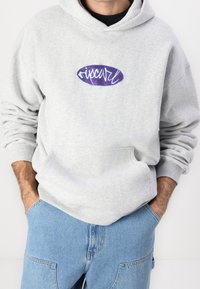 Szara bawełniana bluza z kapturem z fioletowym haftowanym logo na piersi, z kieszenią kangurka i ściągaczami na mankietach. Luźny krój.