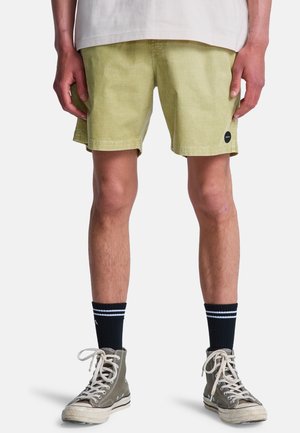 Person trägt blassgrüne Shorts, schwarze Sportsocken mit weißen Streifen und graue High-Top-Sneaker, steht vor einem weißen Hintergrund.
