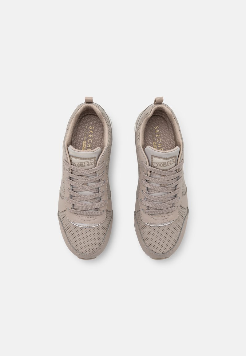 Zapatillas deportivas beige con paneles de malla y puntera redonda, que presentan cordones grises y un tirador en el talón. Logo de Skechers en la lengüeta.