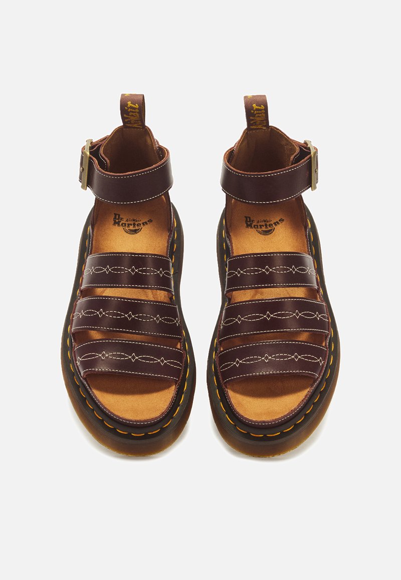 Pair of dark brown leather sandals with three front straps, ankle straps with buckles, and thick black soles with yellow stitching.