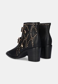 Botas negras hasta el tobillo con un acabado texturizado, que presentan tachuelas en tono dorado y dos hebillas. Diseño de tacón cuadrado, mostrando patrones de costura detallados.