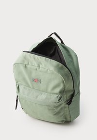 Mochila de tela verde con una textura suave, forma redondeada, cierre de cremallera, bolsillo frontal y una etiqueta de logo en el centro superior.