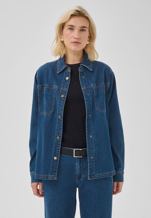 Femme aux cheveux blonds mi-longs portant une veste en jean bleue, un t-shirt noir, un jean bleu et une ceinture noire, debout devant un fond uni.