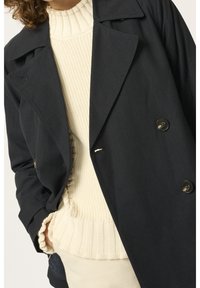Femme portant un manteau noir croisé au-dessus d'un pull en maille côtelée crème avec des poignets et un ourlet à volants, mains dans les poches.