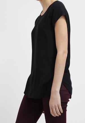 Vrouw draagt een zwart kort mouwtjes shirt met een ronde hals en donkere bordeauxrode broek, gezien van opzij tegen een effen achtergrond.