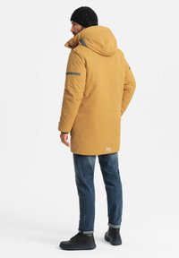 Geel geïsoleerde parka met een capuchon, grijze reflecterende strepen en een logopatch aan de zoom, gedragen over een donkere spijkerbroek en zwarte laarzen.