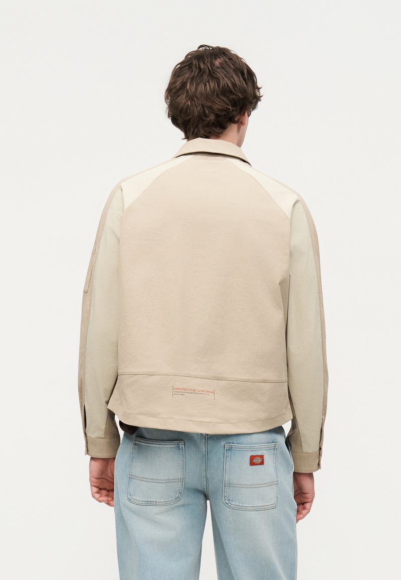 Homme portant une veste beige avec un texte près de l'ourlet et un jean bleu clair avec deux poches arrière et un écusson logo marron, de dos sur un fond uni.
