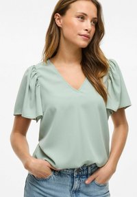 Menta zelena bluza s V-izrezom, kratkim napuhanim rukavima i glatkom teksturom. Uparena s svijetloplavim trapericama.