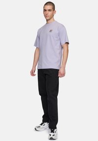 Camiseta corta de manga en color lila con un parche de logo cuadrado. Combinada con jeans negros y zapatillas blancas, de pie sobre un fondo uniforme.