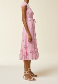 Rosa Blumenkleid mit kurzen, rüschengeschmückten Ärmeln, einem taillierten Oberteil und einem Bindeband in der Taille. Ausgestellte, rüschengeschmückte Saumlinie. Kombiniert mit weißen, schnürbaren Sandalen.