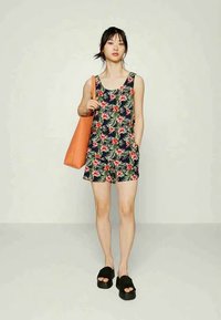 Macacão com estampado floral em azul-marinho, com folhas verdes e flores cor-de-rosa, apresentando um decote em forma de urna, bolsos e combinado com uma bolsa de mão laranja brilhante.