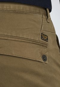 Khaki bukser med en glat tekstur, forstærkede sømme og et lille sort mærke på lommen med gul og hvid tekst.