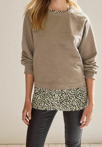 Felpa in taupe chiaro con maniche lunghe e colletto a coste, sovrapposta a una base crema con macchie leopardate nere; abbinata a leggings neri.
