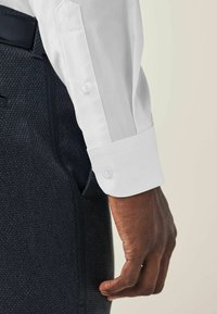 Weißes Hemd mit knöpfbaren Manschetten, kombiniert mit einem strukturierten dunklen Stoff. Nahaufnahme des Handgelenks und der Hand, die das Knopfdetail zeigt.