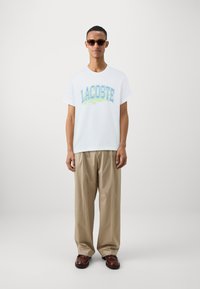 Balta īsām piedurknēm T-kreklā ar krāsainu 'LACOSTE' logo, apvienota ar bēšām platām biksēm un brūniem ādas apaviem.