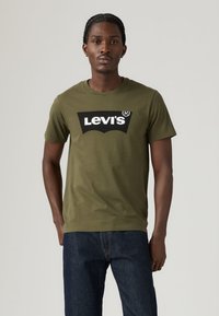 T-shirt de manga curta verde azeitona, feita de algodão, com um grande logótipo preto da Levi's no peito, combinada com jeans azul escuro.