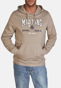 Beżowa bluzka z kapturem z przednią kieszenią typu kangur, sznurkiem do regulacji kaptura i logo "MUSTANG" w granatowych akcentach; zestawiona z jasnoniebieskimi dżinsami.