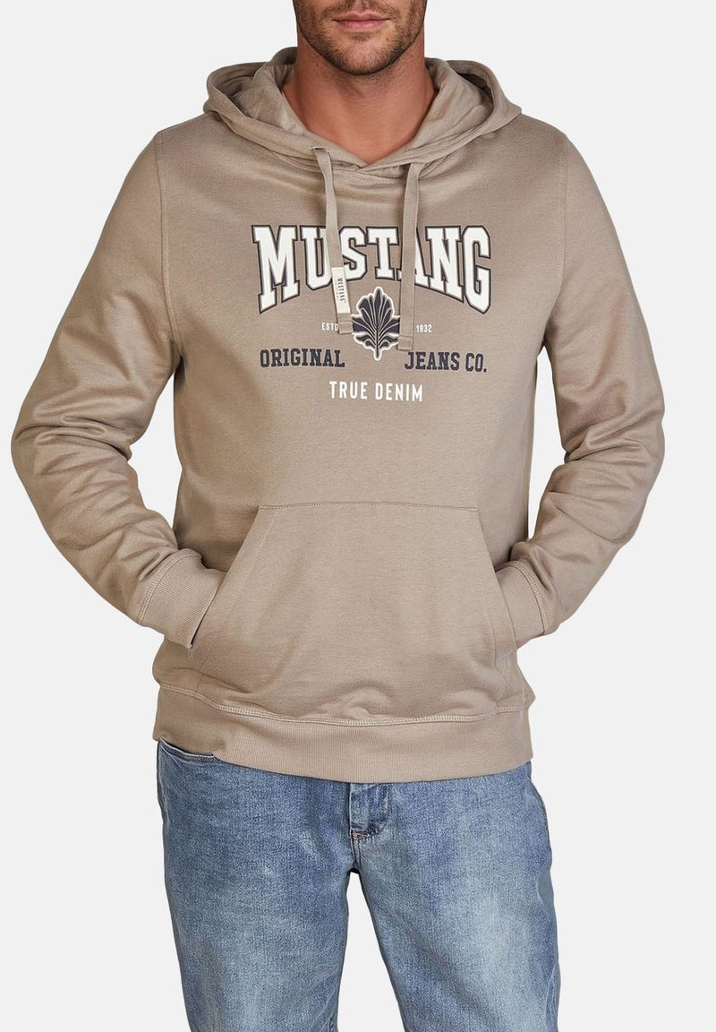 Beżowa bluzka z kapturem z przednią kieszenią typu kangur, sznurkiem do regulacji kaptura i logo "MUSTANG" w granatowych akcentach; zestawiona z jasnoniebieskimi dżinsami.