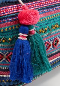 Des pompons multicolores avec des fils roses, bleus et verts, rehaussés de perles, attachés à un sac en textile tissé aux motifs complexes.