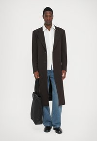 Abrigo largo marrón con solapas anchas, diseño de un solo botón, llevado sobre una camisa blanca, combinado con vaqueros azules de Corte acampanado y un bolso tote negro.