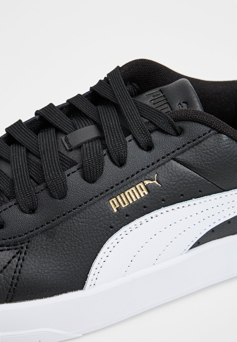 Baskets en cuir noir avec des accents blancs et le logo doré de Puma. Présente des lacets noirs et une surface texturée. Design bas avec une semelle en caoutchouc.