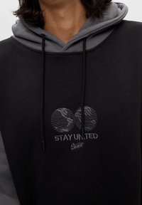 Sudadera negra con capucha gris oscuro, que presenta un gráfico bordado de un globo y el texto "MANTENTE UNIDO". Bolsillo canguro forrado y cuerdas ajustables.