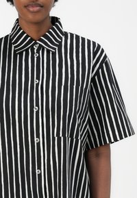 Čierno-biely pruhovaný tričko vyrobené z bavlny. Obsahuje klasický golier, kratšie rukávy, predné zapínanie na gombíky a vrecko na hrudi.