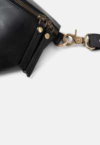 Black leather bag corner with double zipper pulls and brass snap hook clasp on strap against white background.