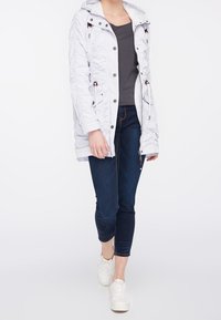 Veste matelassée gris clair avec capuche, taille à cordon et boutons décoratifs. Associée à un jean foncé et à des baskets blanches.