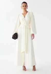 Vestido largo color crema con escote en V profundo, cintura ceñida y mangas fluidas. Acompañado de un clutch negro texturizado y tacones beige con tiras.