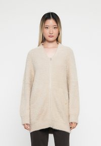 Beige ritsvest gemaakt van zacht, textuur materiaal met een losse pasvorm, lange mouwen en twee voorkeurszakken.