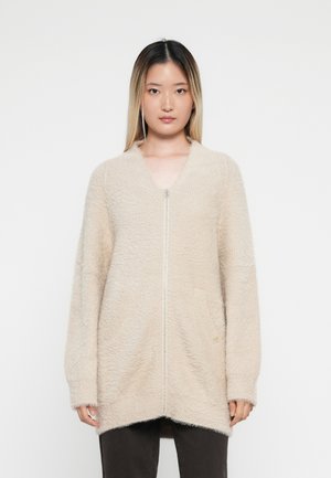 Beige ritsvest gemaakt van zacht, textuur materiaal met een losse pasvorm, lange mouwen en twee voorkeurszakken.