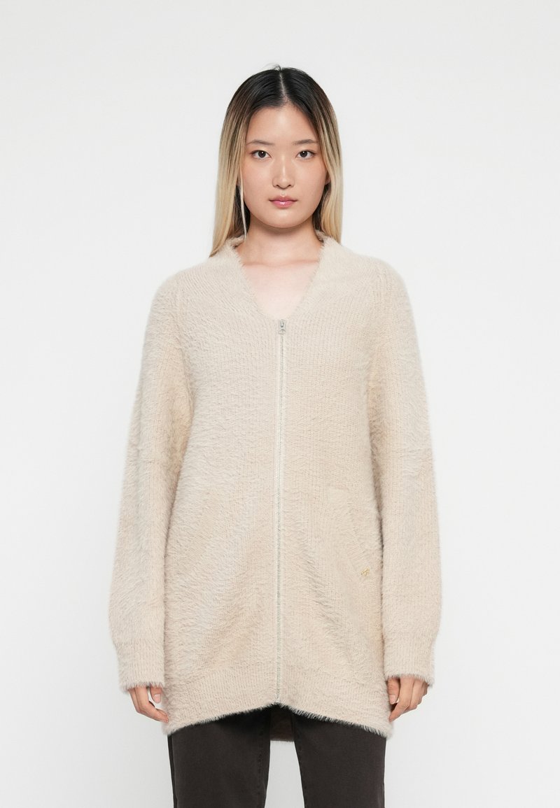 Beige ritsvest gemaakt van zacht, textuur materiaal met een losse pasvorm, lange mouwen en twee voorkeurszakken.
