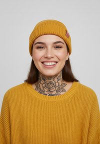 Mustard yellow knitted beanie with a ribbed texture, worn with a matching sweater. Notable floral tattoo on neck. Neutral background.