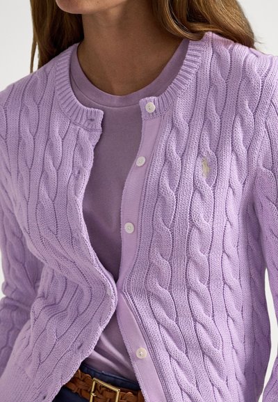 Καρδιγκάν με πλέξη καλώδιο σε λεβαντρί απόχρωση, με στρογγυλή λαιμόκοψη και λευκά κουμπιά, φορεμένο πάνω από ένα μονόχρωμο λεβαντρί πουκάμισο. Η υφή του υφάσματος προσθέτει βάθος.