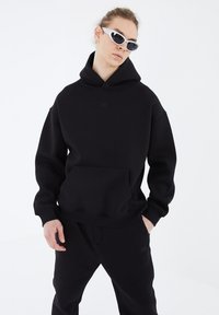 Black hooded sweatshirt made of textured fabric with a kangaroo pocket and subtle front branding. Paired with matching pants and sunglasses.