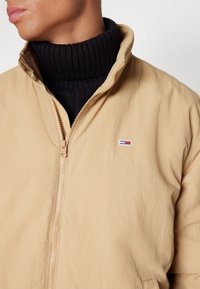 Béžová zipová bunda s výstřihem ke krku, vyrobená z lehkého materiálu. Má malé logo na hrudi a je doplněná černým žebrovaným rolákem.