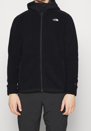 Homme portant une veste polaire noire zippée avec capuche et logo The North Face sur la poitrine, associée à un pantalon noir.