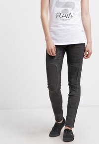 Schwarze enge Jeans mit strukturierten Einsätzen an den Knien, mit einem abgenutzten Finish und schlanker Passform. Kombiniert mit einem weißen Grafik-Top und Turnschuhen.