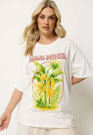 Mujer rubia con camiseta blanca de gran tamaño con ilustración colorida de planta de plátano y texto "Banana Botanica" sobre fondo liso.
