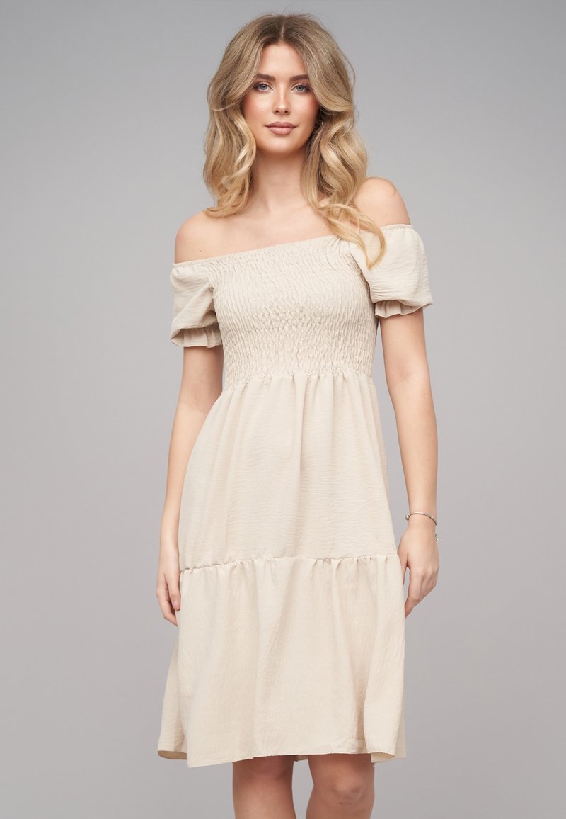 Femme aux longs cheveux blonds ondulés portant une robe beige épaules dénudées, longueur genou, avec des manches ballon, debout devant un fond gris uni.