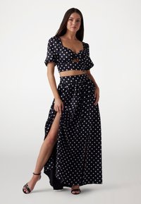 Conjunto de dos piezas negro con lunares que incluye un crop top ajustado con mangas abullonadas y una falda maxi de alta abertura, fabricado con una tela suave y brillante.