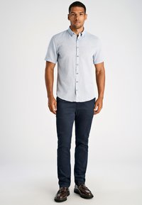 Koszula z krótkim rękawem w jasnoniebieskim kolorze, z guzikami i teksturowanym materiałem, zestawiona z ciemnoniebieskimi chinosami i brązowymi eleganckimi butami. Model stoi prosto.