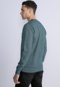 Türkiser Fleece-Pullover mit gerippten Bündchen und Saum, ausgestattet mit einem Rundhalsausschnitt und einer strukturierten Oberfläche, kombiniert mit schwarzen Jeans.