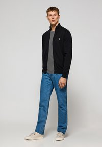 Zwarte zip-up jack over een grijze t-shirt, gecombineerd met lichtblauwe jeans en beige sneakers. Het jack heeft een klein logo op de borst.